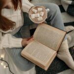 fair skinned, red-haired person sitting cross-legged and reading with a cup of hot chocolate