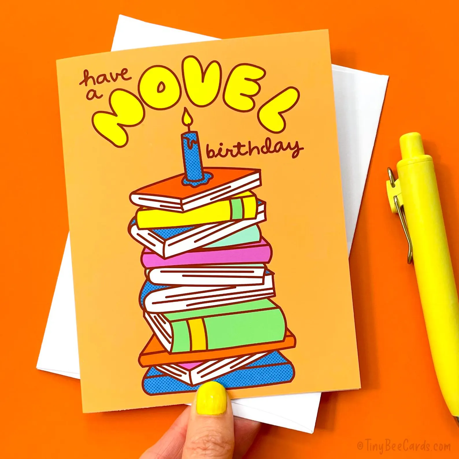 image of a birthday card with a bright orange background. There is a "cake" made of books that says "have a novel birthday." 