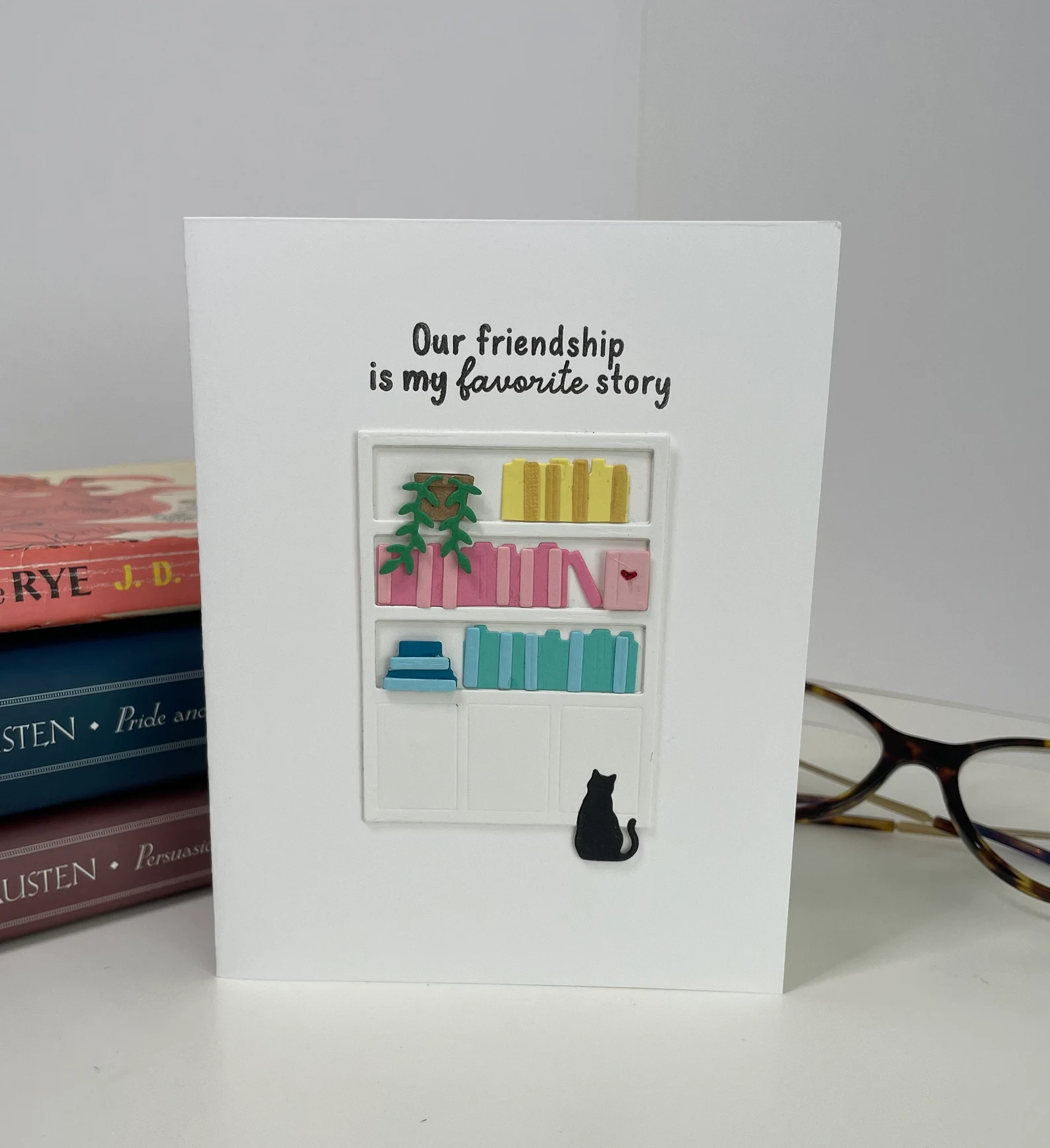 Image of a card that features a small bookshelf and a black cat looking up at the shelf. It reads "Our friendship is my favorite story."