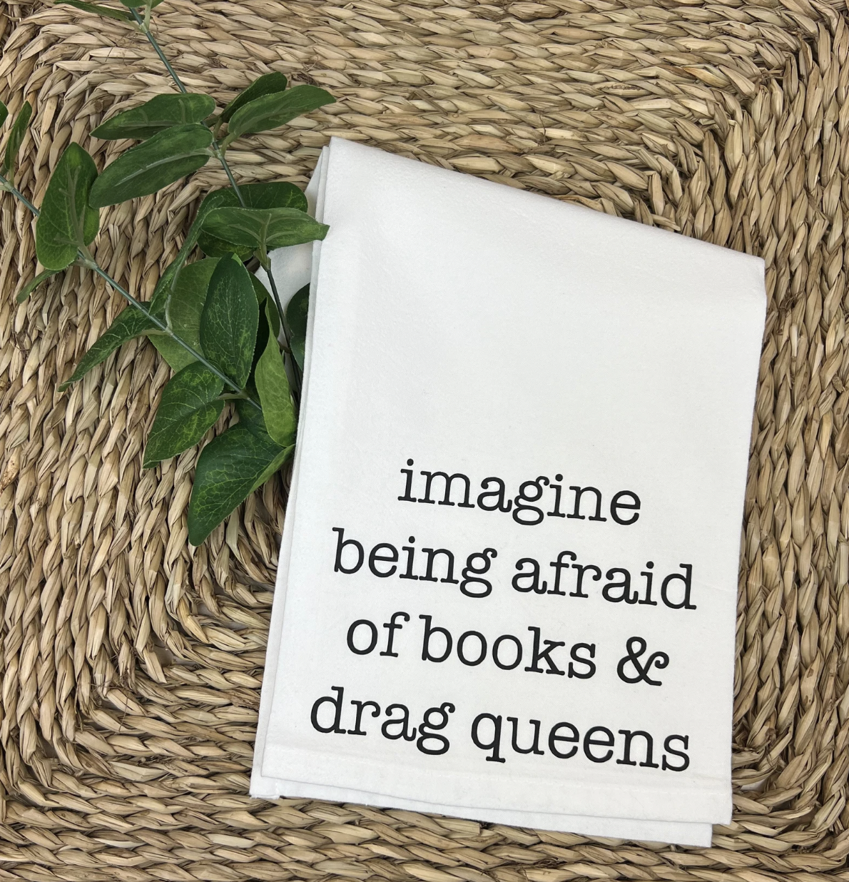 Image of a white tea towel with the phrase "imagine being afraid of books and drag queens" screenprinted in black font