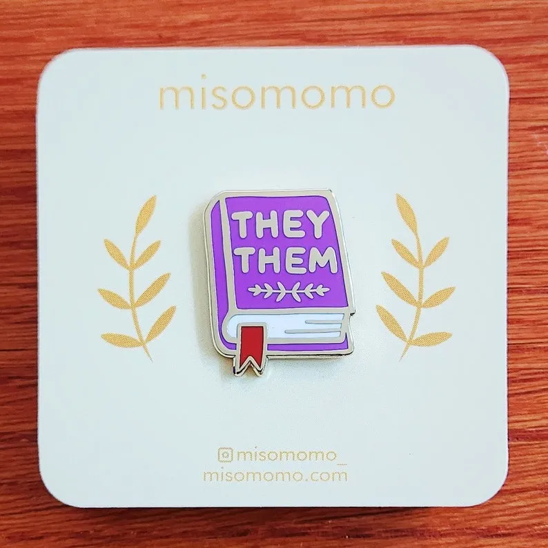 an enamel pin of a book with the text They/Them
