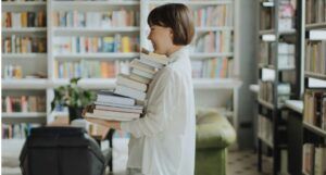 a fair-skinned Asian woman with a short bob holding a big stack of books and laughing