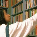 a light brown-skinned woman with dark hair reaching for a book in a used bookstore