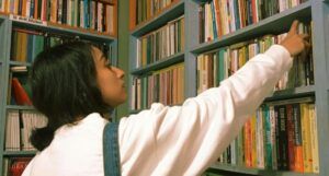 a light brown-skinned woman with dark hair reaching for a book in a used bookstore