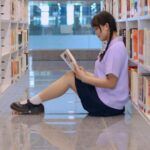 a light-skinned young Asian woman sitting on a bookstore floor reading a book