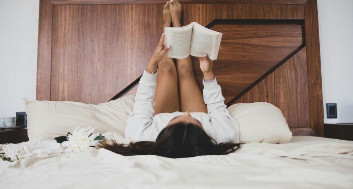 a light-tan skinned young woman reading a book in bed with her feet resting on the head board