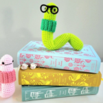 Crocheted amigurumi bookworms with cute little smiles and round glasses sitting atop a stack of books