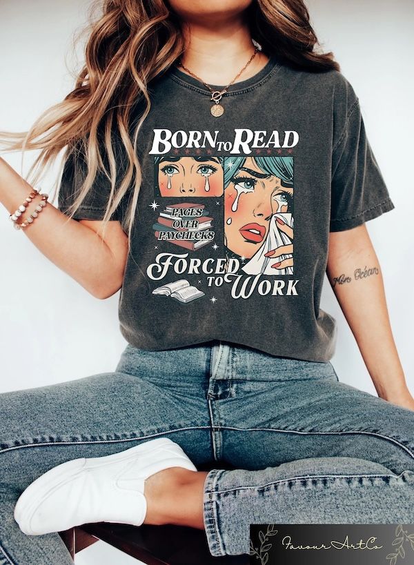 a woman's torso in a dark grey t-shirt containing comics-style graphics of a woman crying bordered by white text that reads "Born to Read, Forced to Work"