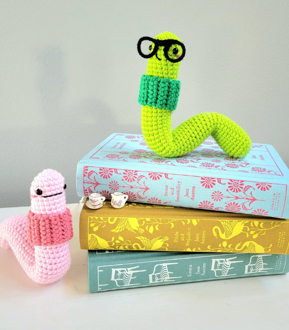 Crocheted amigurumi bookworms with cute little smiles and round glasses sitting atop a stack of books