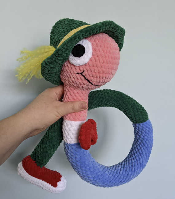 Crocheted stuffie of the character Lowly Worm from Richard Scarry's Busyworld wearing a red bowtie, red shoe, and green hat