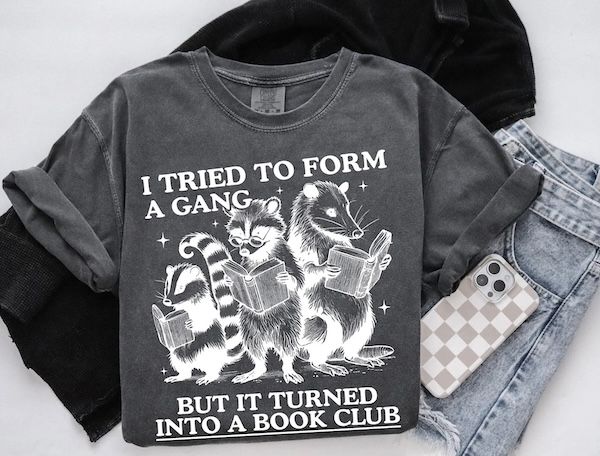 a dark grey t-shirt showing a raccoon, a possum, and a skunk reading books. Text bordering the image reads "I tried to form a gang but it turned into a book club"