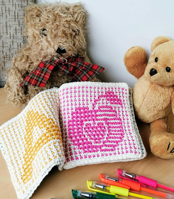 A knitted soft book featuring the capital letter A in yellow and half on an apple in pink lies splayed open against a teddy bear