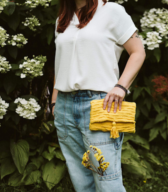 Image of a white woman from the neck down, standing in from of hydrangea bushes, wearing a white blouse and blue jeans while holding a yellow knitted book clutch bag in her hand with a tassle