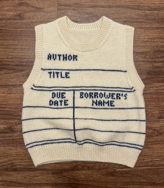 A white knitted vest designed to look like a library checkout card with space for the author, title, due date, and borrower's name laying against a wooden background