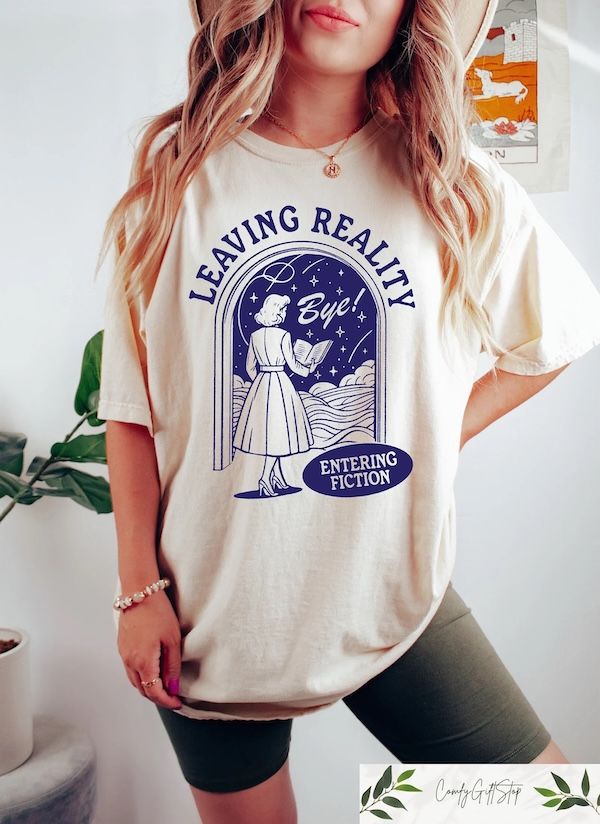 torso of a woman wearing a cream t-shirt with a graphic of a women in an a-line dress holding a book while walking through a doorway. Bordering the graphic is text that reads "leaving reality, entering fiction"