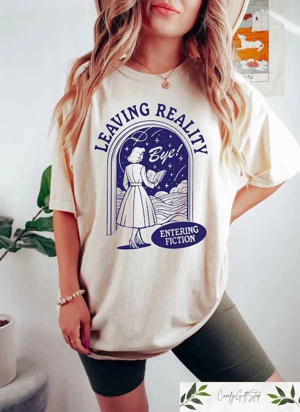 torso of a woman wearing a cream t-shirt with a graphic of a women in an a-line dress holding a book while walking through a doorway. Bordering the graphic is text that reads "leaving reality, entering fiction"
