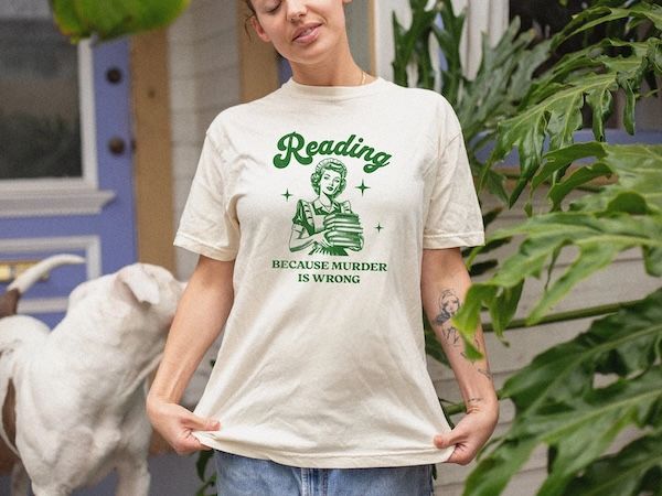 torso of a woman wearing a cream t-shirt with a graphic of a woman holding a stack of books on the front, and green text on top that reads "reading... because murder is wrong"