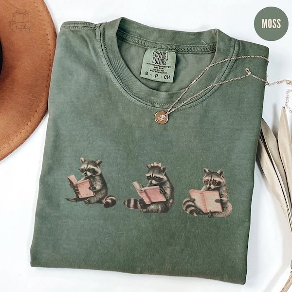 a heather green t-shirt with three seated raccoons on the front, each of them reading a book