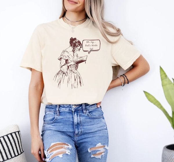 torso of a woman wearing a beige t-shirt with a graphic of a woman in Victorian dress holding a book with a quote bubble hovering to the right that reads "Oh my... that's kinda hot"