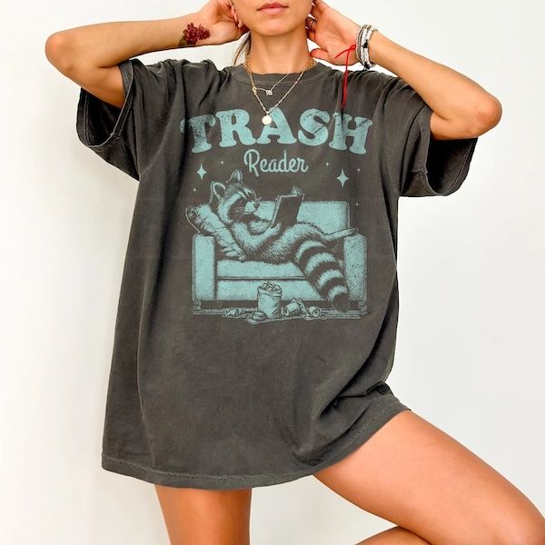 torso of a woman wearing a dark grey shirt with a graphic of a raccoon reading in an armchair on the front. Text above the graphic reads "Trash Reader"