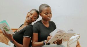 two brown-skinned Black women with short haair sitting back to back and reading books