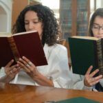 two women of color sitting next to each other and reading books at a desk in a library