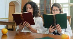 two women of color sitting next to each other and reading books at a desk in a library