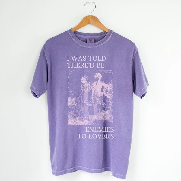 purple t-shirt on a hanger with a graphic of a couple in Regency dress on the front. Text bordering the image reads "I Was Told There'd be Enemies to Lovers"