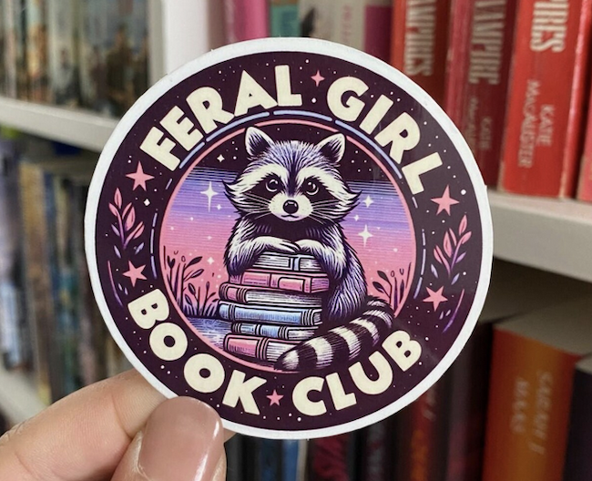 a round sticker illustrated with a raccoon holding a book stack that says feral girl book club