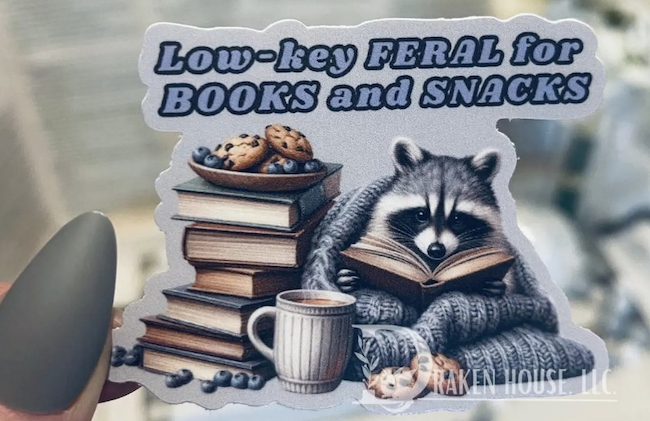 a sticker of an illustrated reacoon with snacks reading a book with text that says low key feral for books and snacks