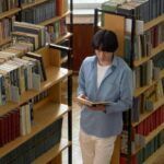 a light-skinned Asian man reading a book in a library