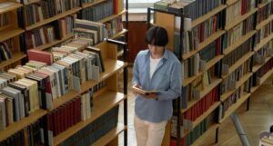 a light-skinned Asian man reading a book in a library