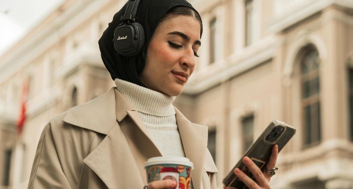 a light tan-skinned woman with a headscarf looking at her phone while wearing headphones