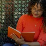 a light tan skinned woman with long, curly hair and an orange shirt reading a book in a wicker chair