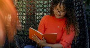 a light tan skinned woman with long, curly hair and an orange shirt reading a book in a wicker chair