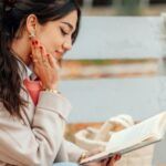 a lightly tan-skinned woman sitting pleasantly reading a book while sitting on a bench (1)