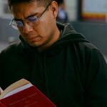 a tan-skinned man with glasses reading a book on a sidewalk