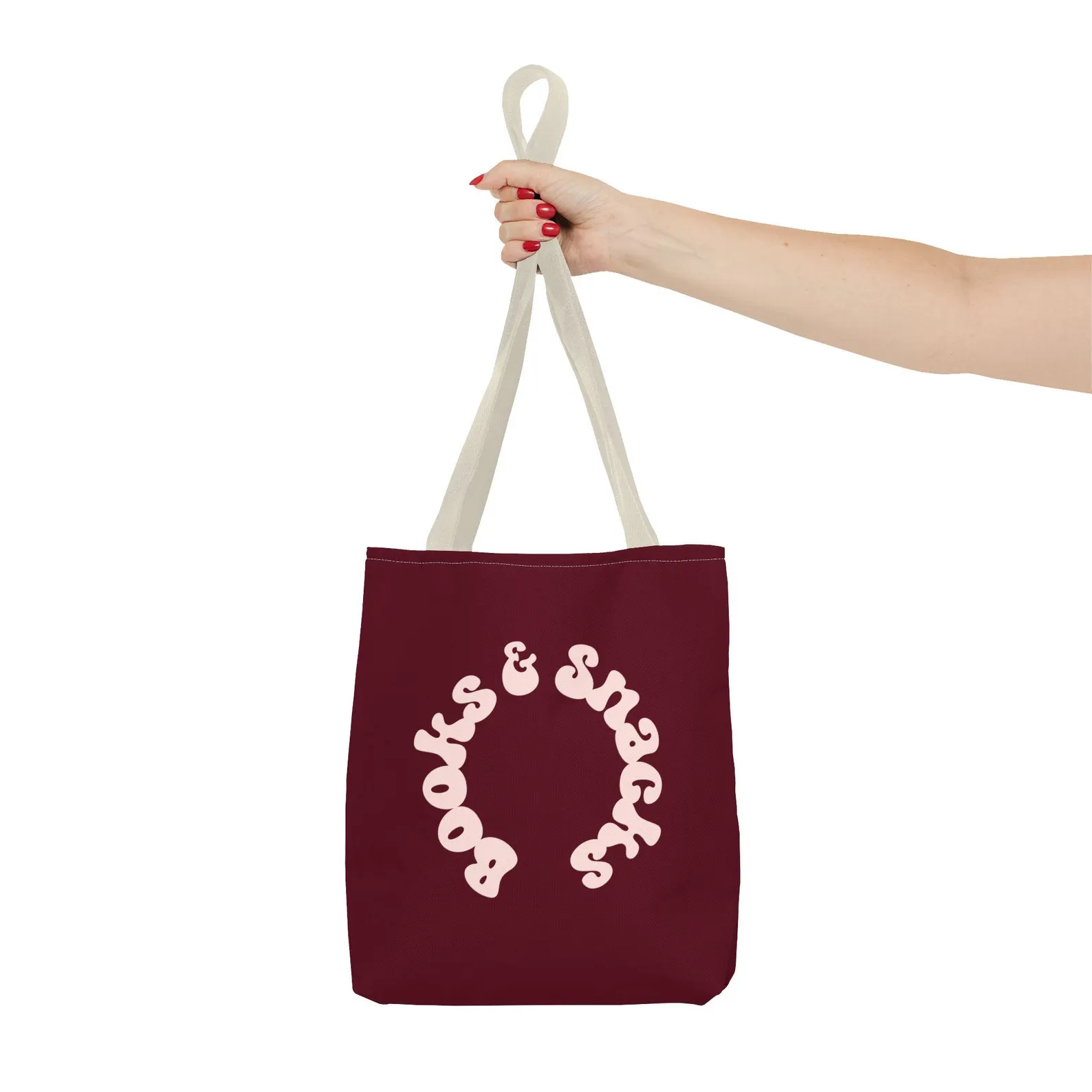 A white hand holding a tote bag. The tote bag is maroon with pink font that reads "books and snacks."