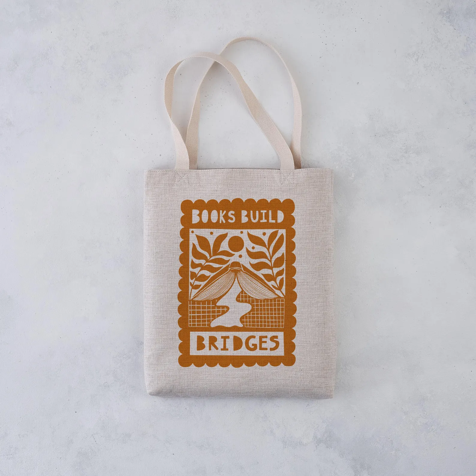 Image of a canvas tote bag. There is an ochre-colored image featuring a book set up to look like a bridge over a river. In white it reads "books build bridges."