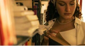 fair-skinned woman with curly hair and a scarf reading a book against a bookshelf