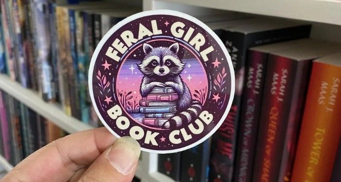 a sticker of a raccoon on a stack of books with the text Feral Girl Book Club
