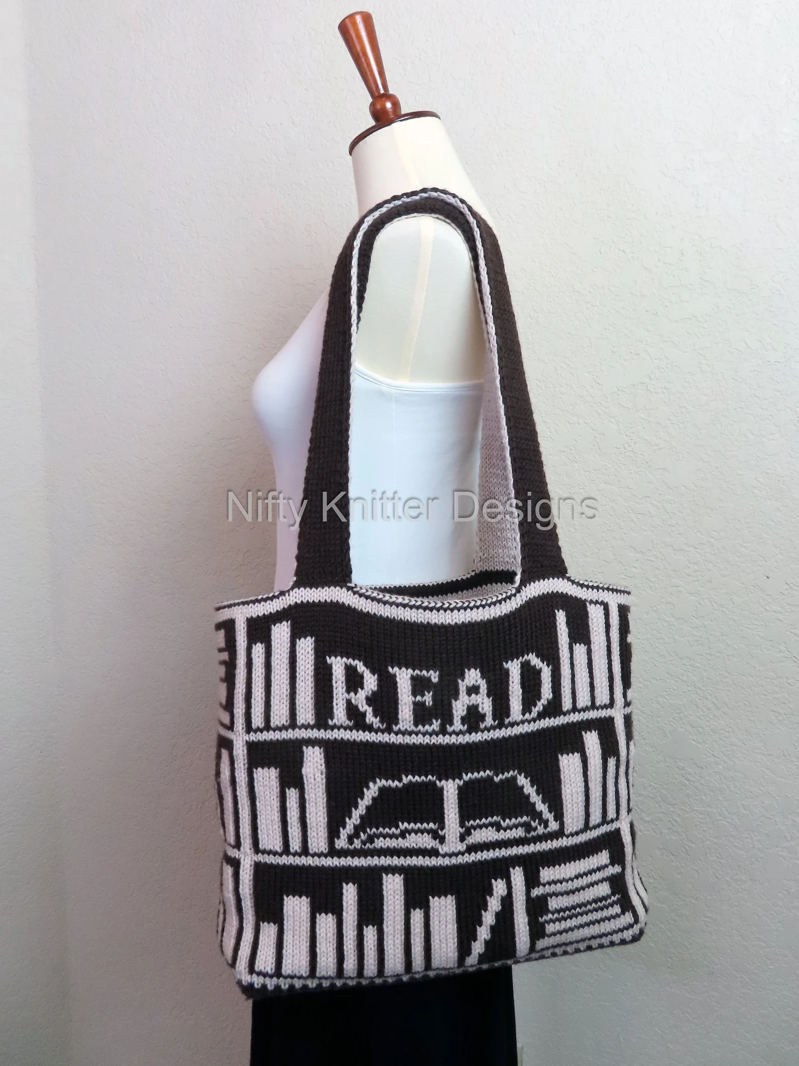 Image of a knit tote bag. It features three bookshelves. On the top bookshelf, it says "read" and on the shelf below, one of the books is open. The tote is black with white books on it. 