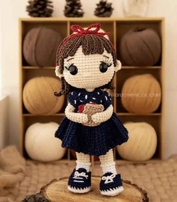 An adorable crocheted doll version of Matilda with a blue dress, red headband, and a book clutched in her arms stands proudly in front of a shelf of yarn. 