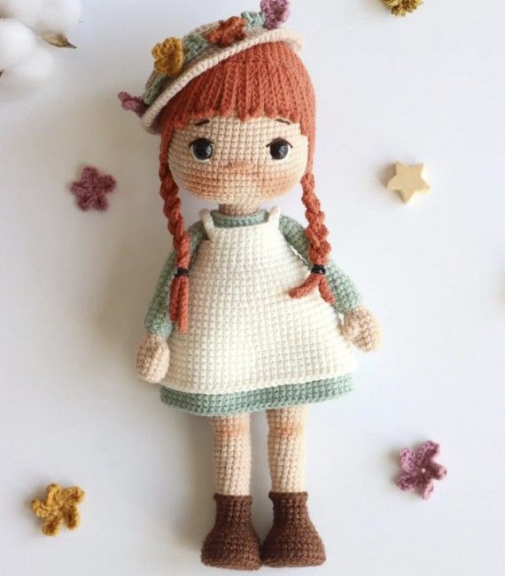 A little crocheted figurine of Anne from Anne of Green Gables wearing a blue dress, white pinafore, and floral hat is rested against a white background with little crocheted flowers. Her red hair is in two braids. 