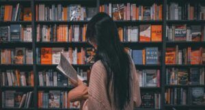 asian person reading a book in front of a filled bookshelf in a book store
