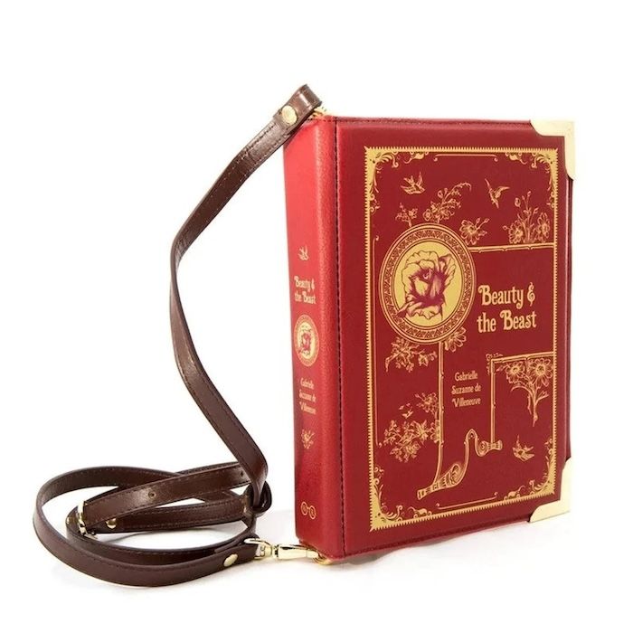 a bag in the shape of a book with a brown leather cross-body strap. The book is a red and gold cover of Beauty and the Beast