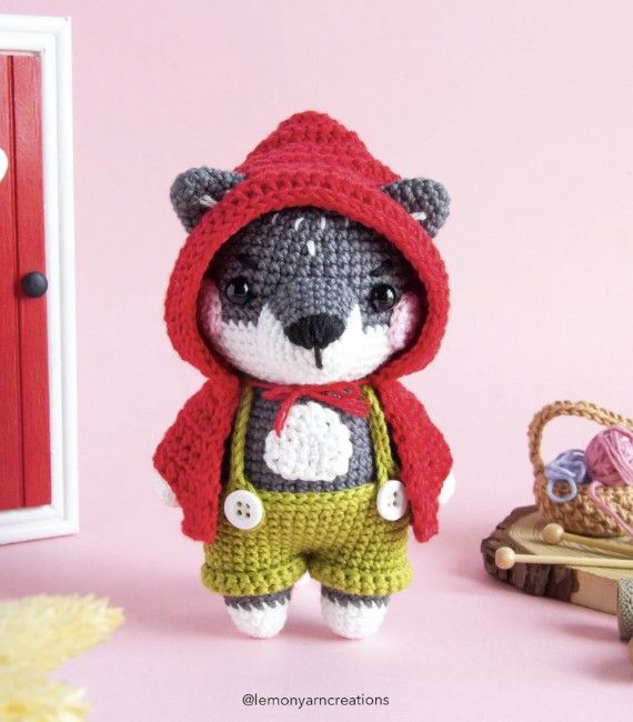 A cute gray wolf amigurumi in green suspendered pants and a red hood stands against a fun pink background. 