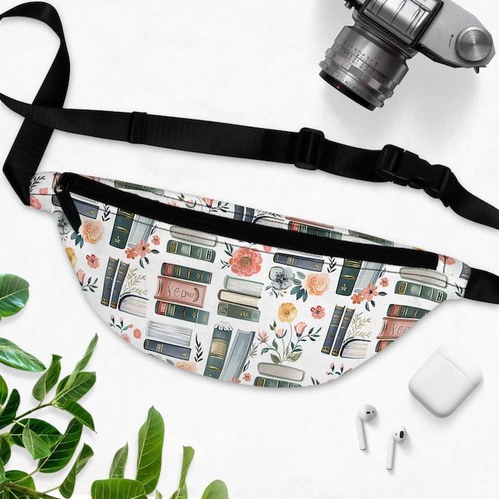 fanny pack with black straps and zipper. the fabric is white with a print of assorted book spines and flowers