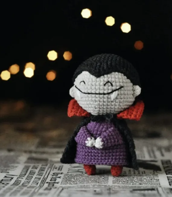 A little crocheted figurine of Dracula with an oversized grey head, purple shirt, and black and red cape boasts a large, fanged smile and happily closed eyes in this picture.