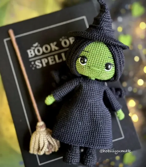 A crocheted doll of Elphaba from the book / musical Wicked is posed on top of a book of spells alongside a broom. She has green skin, black hair, and is wearing a black dress and black hat.